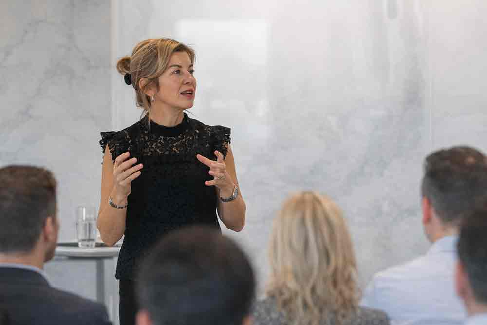 Dr. Julie Gowthorpe speaking to an audience at a professional event, gesturing with her hands while presenting in a bright, modern room.